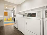 Coin Operated Washing and Drying Machines and Folding Table with Open Doorway to Yellow Chairs in Lounge Area  