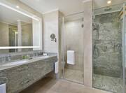 Hotel Suite Bathroom with Long Walk-in Shower, Illuminated Mirror above Vanity with Two Sinks all in a Light Gray Granite
