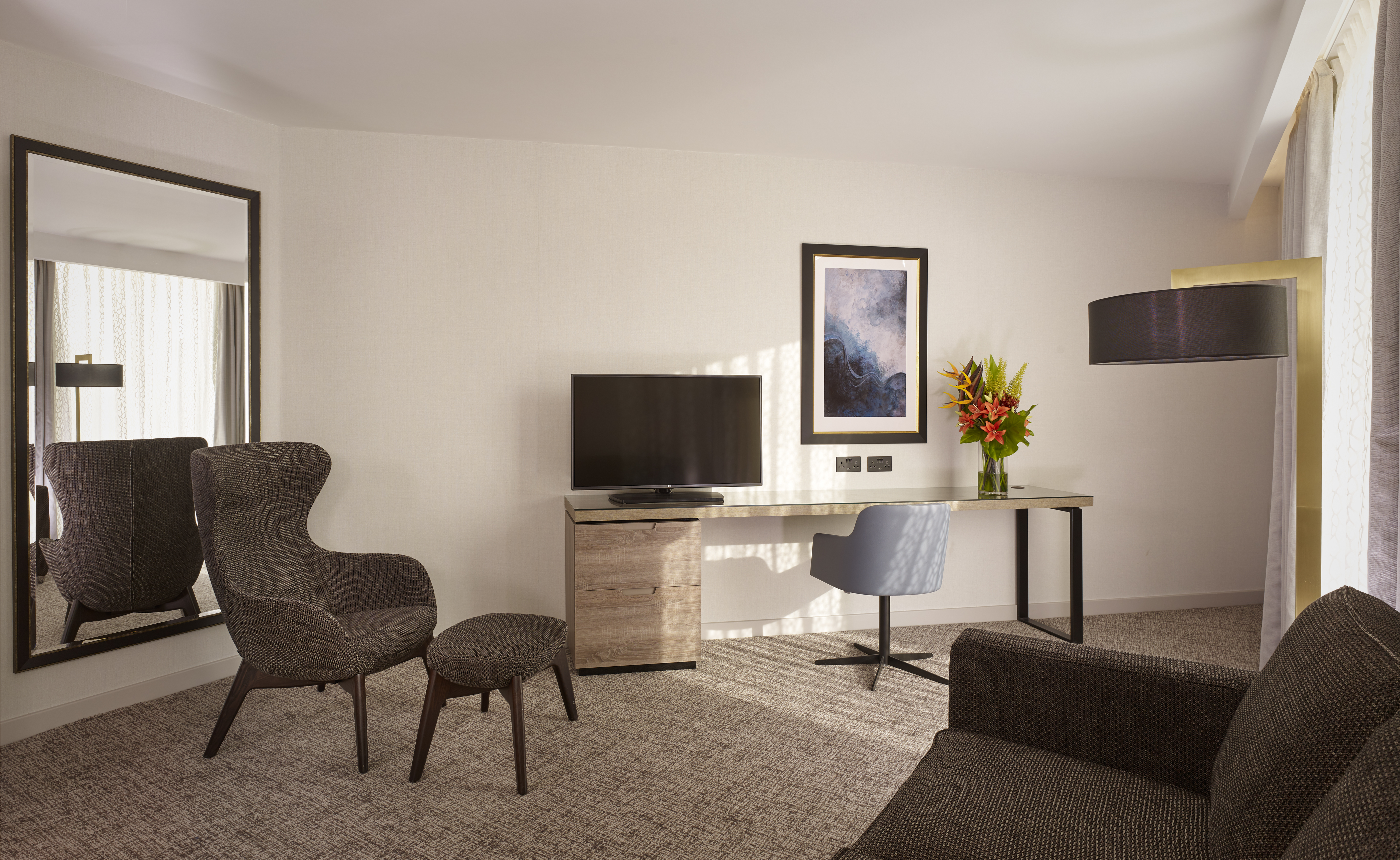 Living Area of Guest Suite with Large Windows and Natural Light coming in, a Floor to Ceiling Mirror, HDTV, Gray Chair for Natural Wood Desk