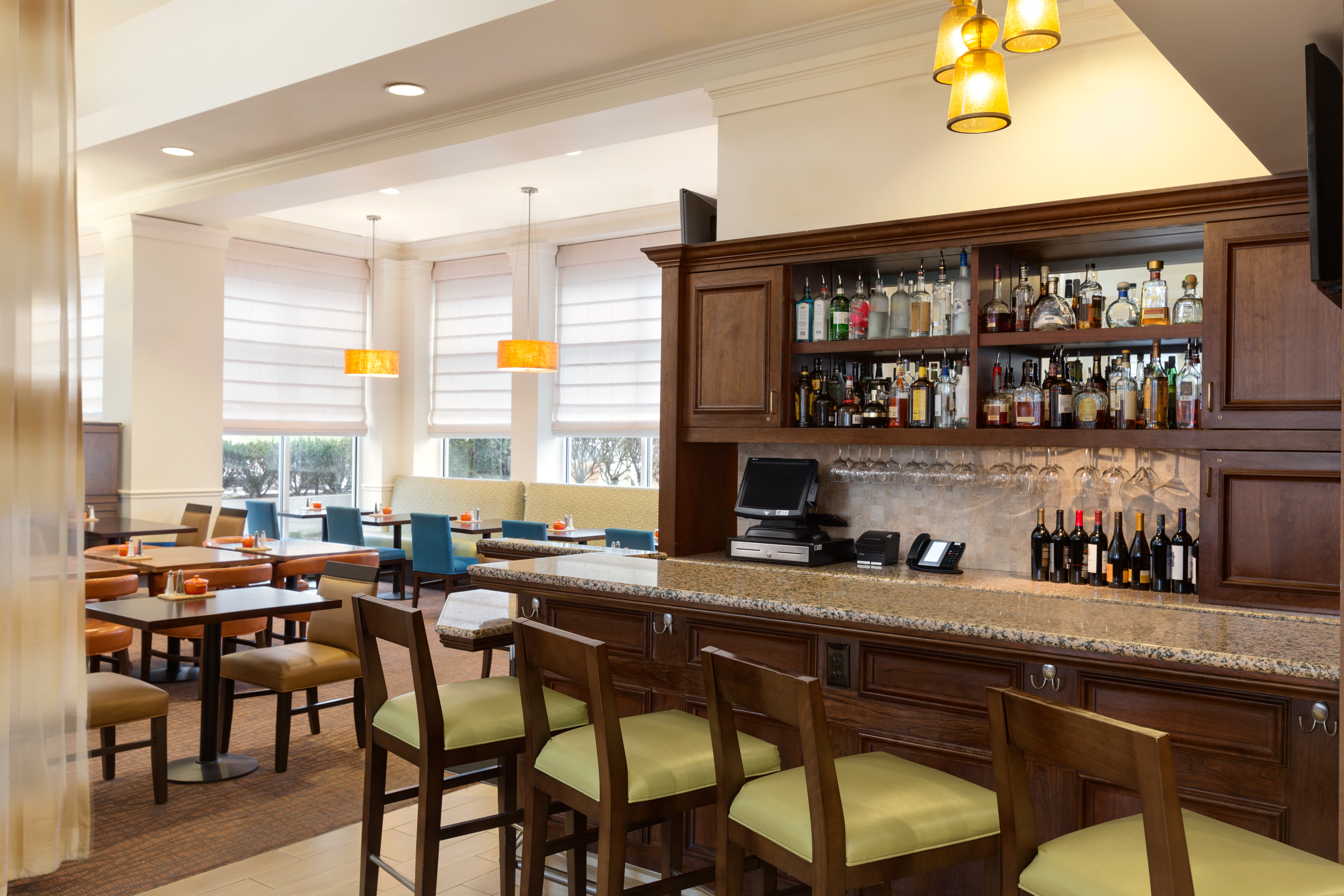 Fully Stocked Bar With Angled Green Bar Chairs and Table Seating by Windows in Background