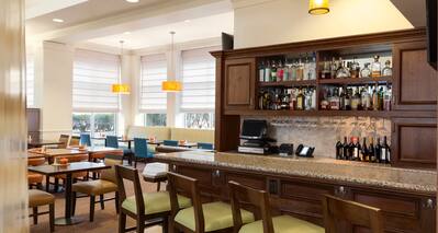Fully Stocked Bar With Angled Green Bar Chairs and Table Seating by Windows in Background