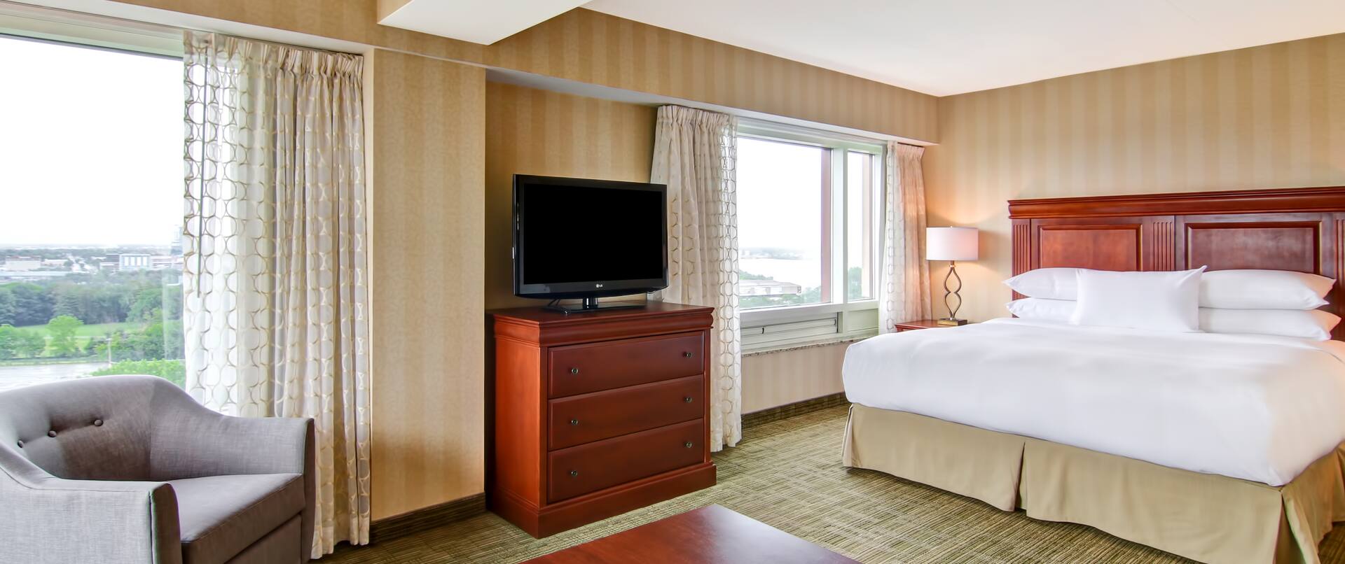 Suite with king bed, work desk, soft chair, TV, and windows with outdoor view