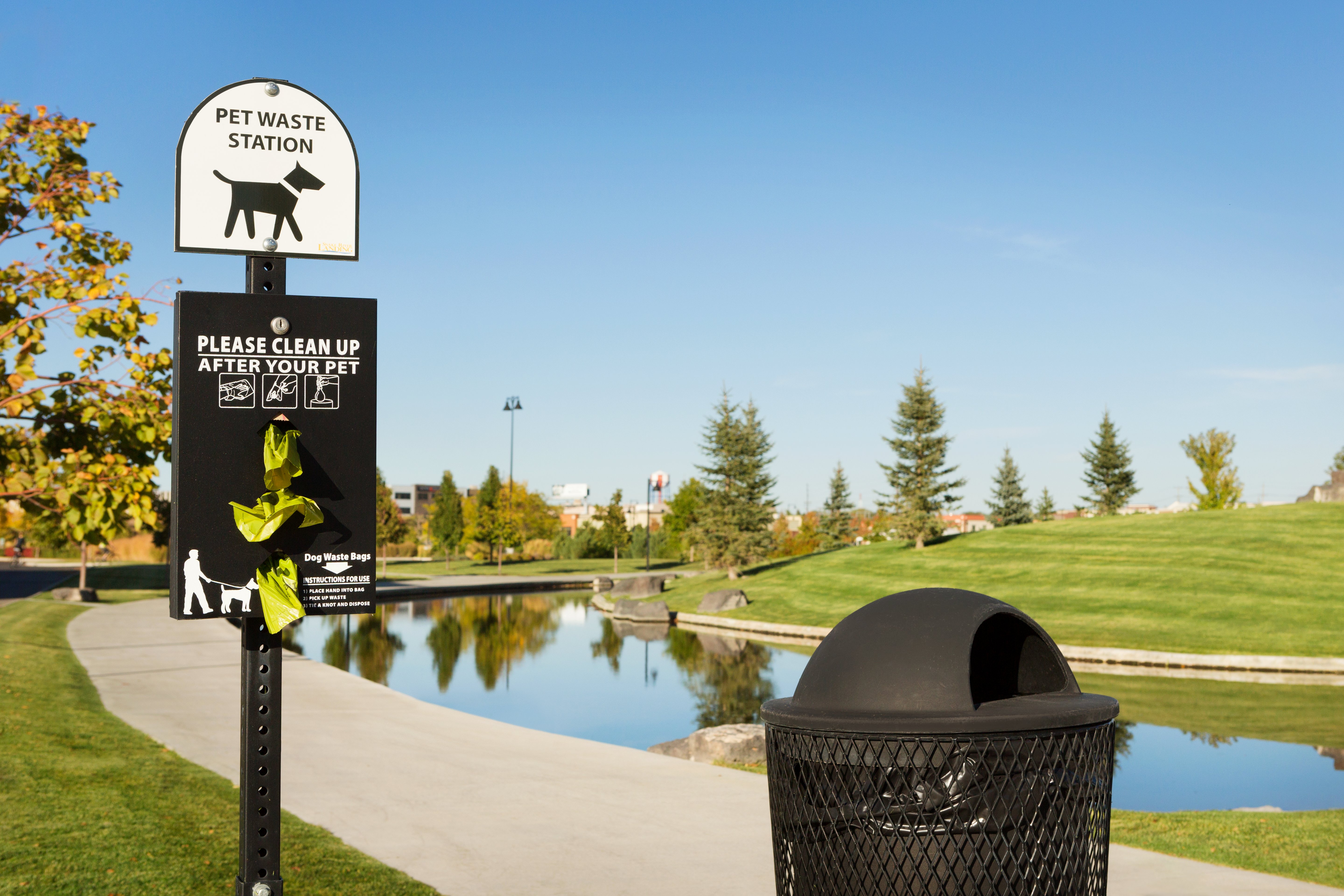 Pet Waste Station Sign, Waste Bag Dispenser, and Black Trash Can in Grassy Pet Area by Water