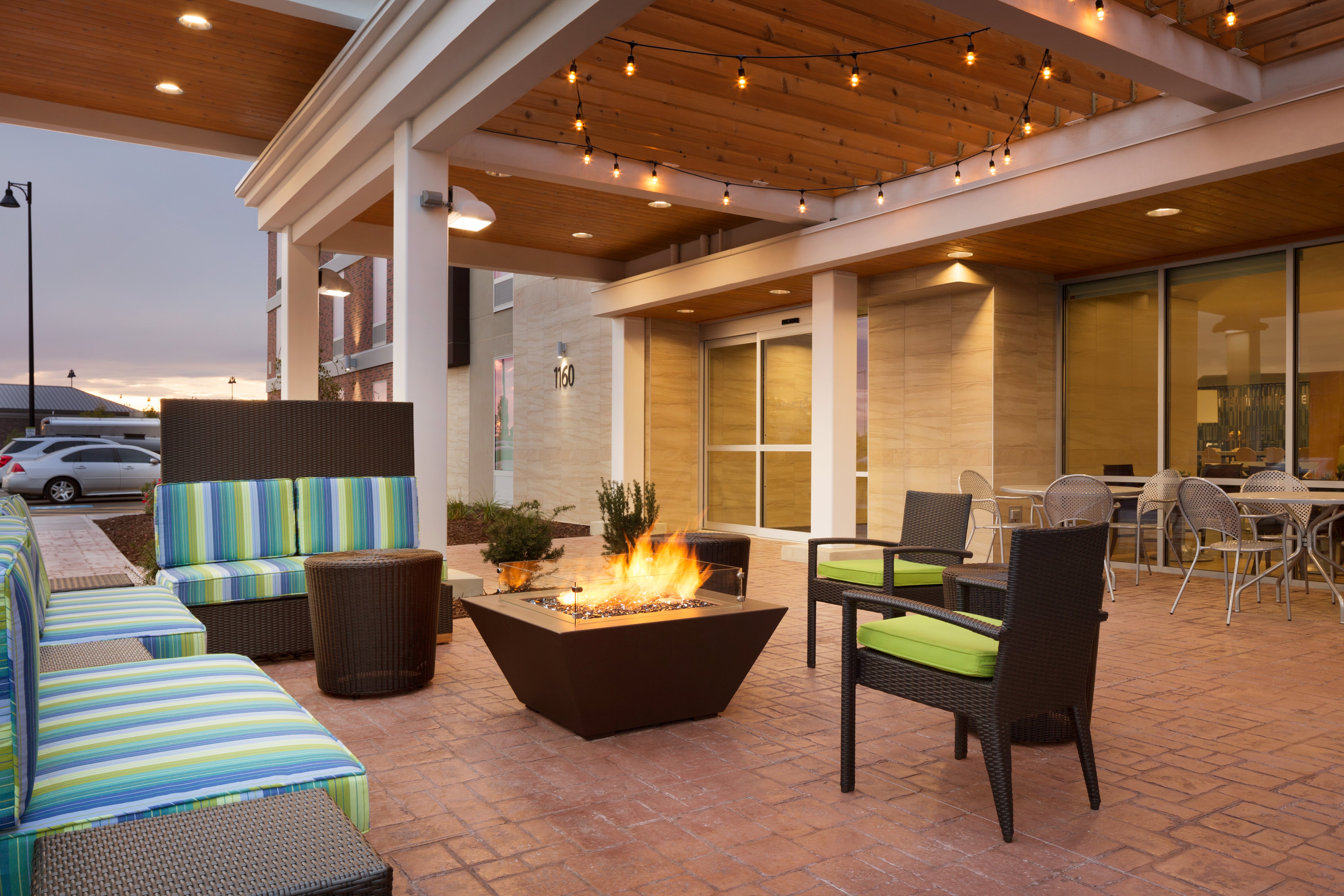 Illuminated Outdoor Lounge Area With Soft Seating Around Fire Pit, Additional Seating by Building and View of Guest Cars on Parking Lot at Dusk