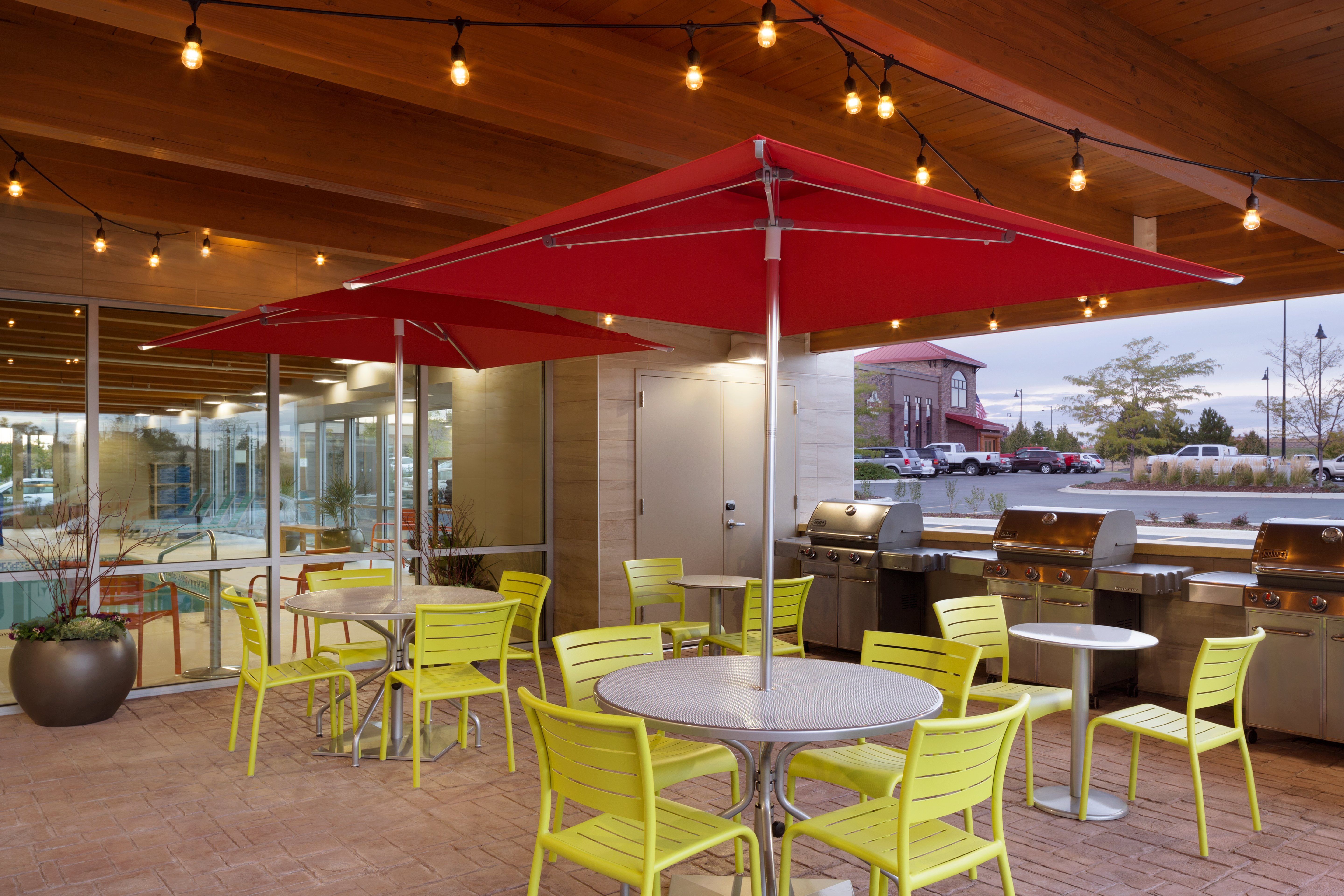 Illuminated Outdoor Patio With Green Chairs, Red Umbrellas Tables, and Three Barbecue Grills at Dusk 