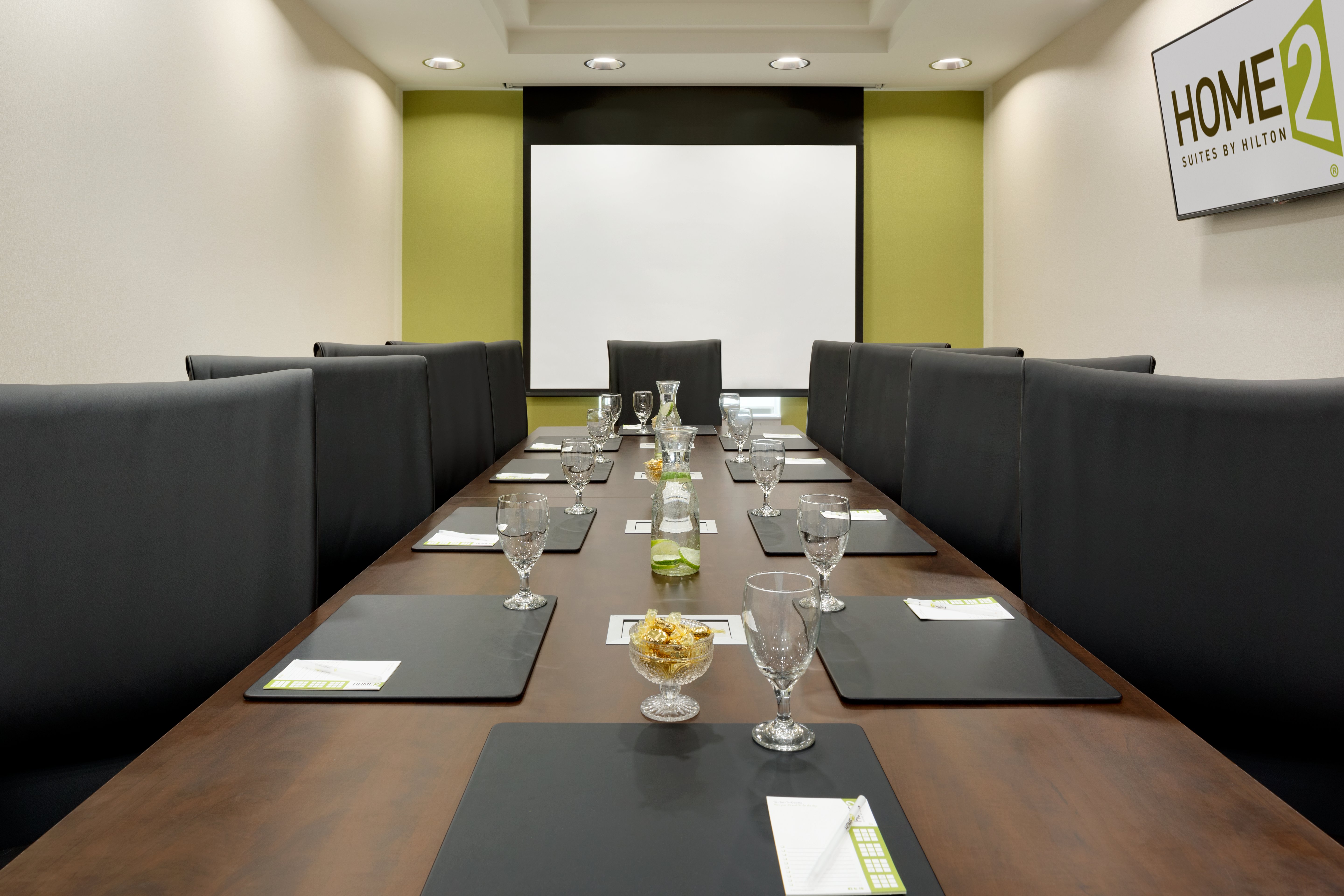 Black Chairs Around Boardroom Table With Presentation Screen on Green Wall and TV