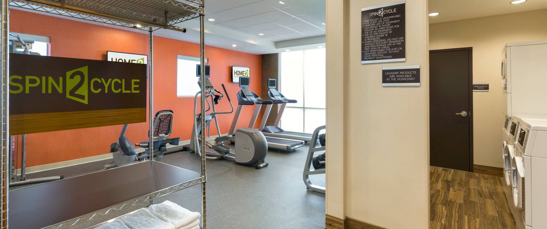 Spin2 Cycle Fitness Center With Cardio Equipment by Window, Towel Rack, and Opening to View of Coin Operated Washing and Drying Machines in Laundry Area