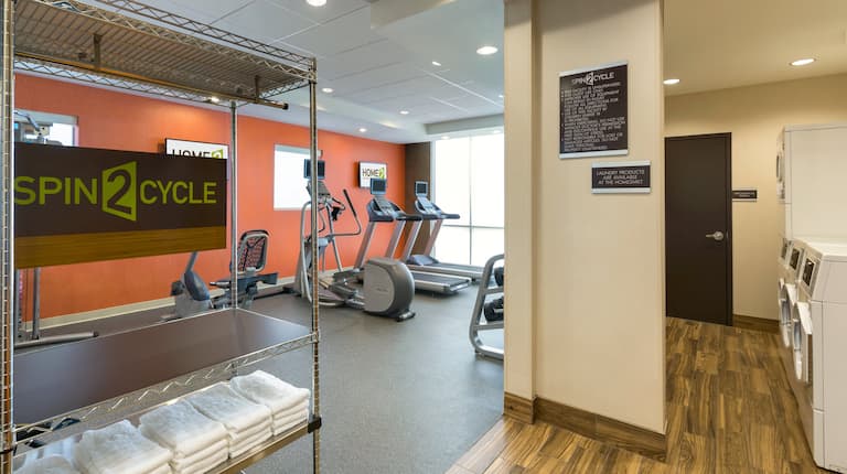 Spin2 Cycle Fitness Center With Cardio Equipment by Window, Towel Rack, and Opening to View of Coin Operated Washing and Drying Machines in Laundry Area