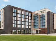 Modern Homewood Suites hotel exterior with chic grey brick, wood paneling, american flag, pristine green lawn, and bright blue sky.