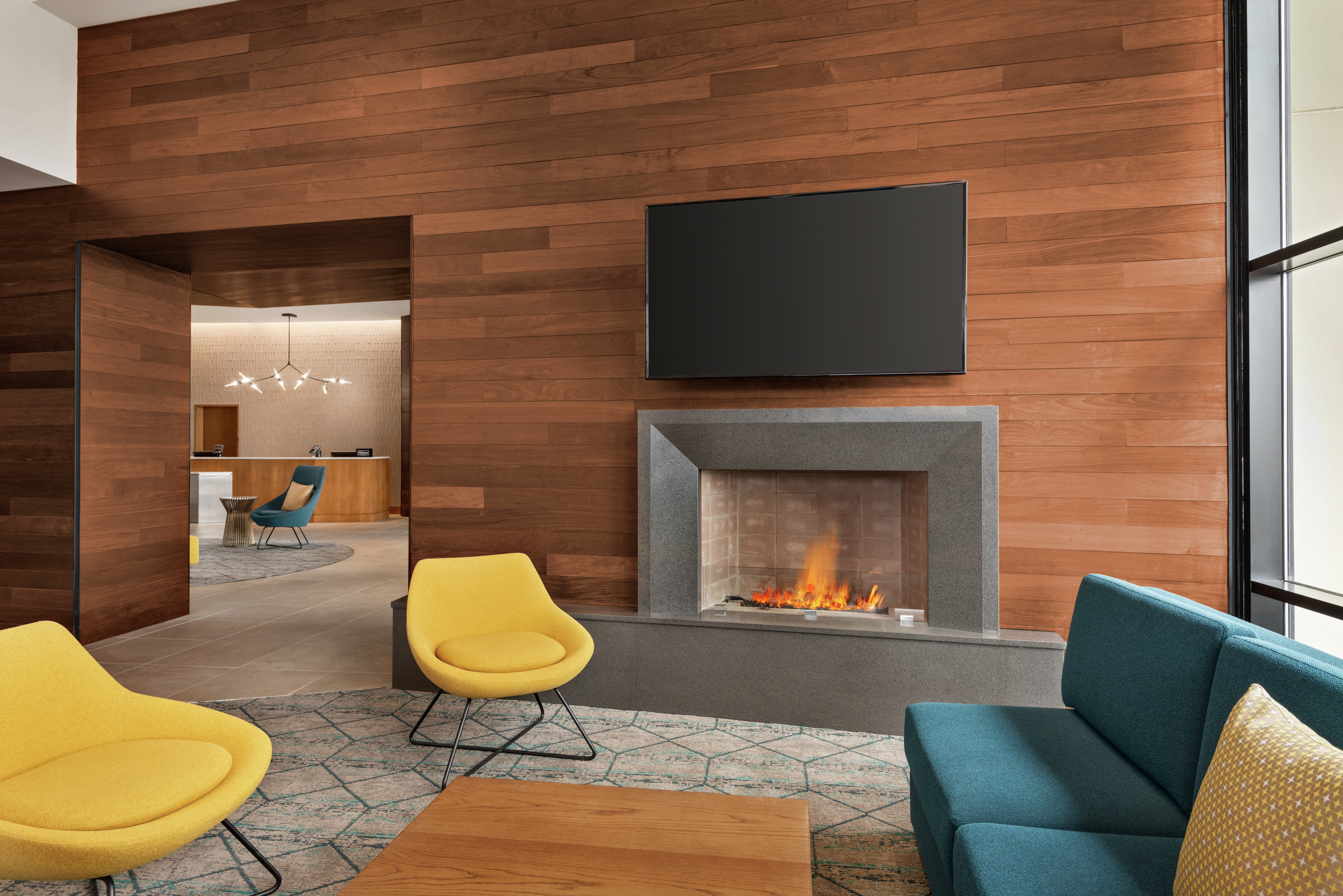 Bright lobby lounge area with stylish furniture, floor to ceiling windows, wood paneled walls, fireplace, and TV.