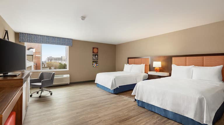 Bright accessible guestroom featuring TV, work desk, two comfortable queen beds, and city outside view.