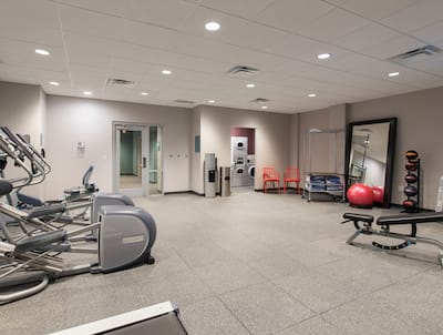 Spin2Cycle Fitness Center With Cardio Equipment, Glass Door Entry, Water Cooler, Open Doorway to Laundry Room, Two Red Chairs, Towel Station, Red Exercise Ball in Front of Large Mirror, Weight Balls, Free Weights, and Weight Bench