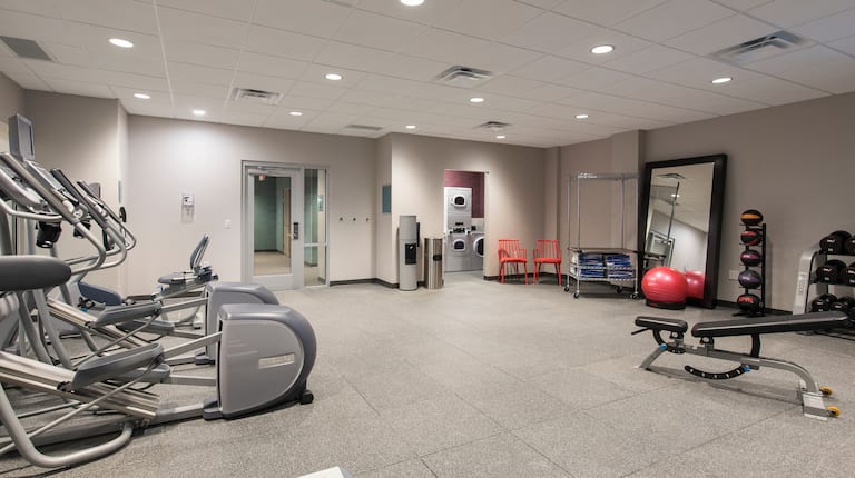 Spin2Cycle Fitness Center With Cardio Equipment, Glass Door Entry, Water Cooler, Open Doorway to Laundry Room, Two Red Chairs, Towel Station, Red Exercise Ball in Front of Large Mirror, Weight Balls, Free Weights, and Weight Bench