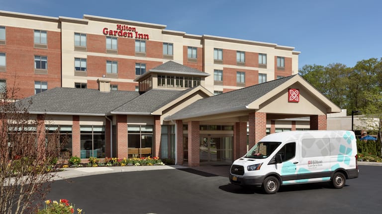 Welcoming Hilton Garden Inn hotel exterior featuring beautiful landscaping, blue sky, and convenient shuttle bus.
