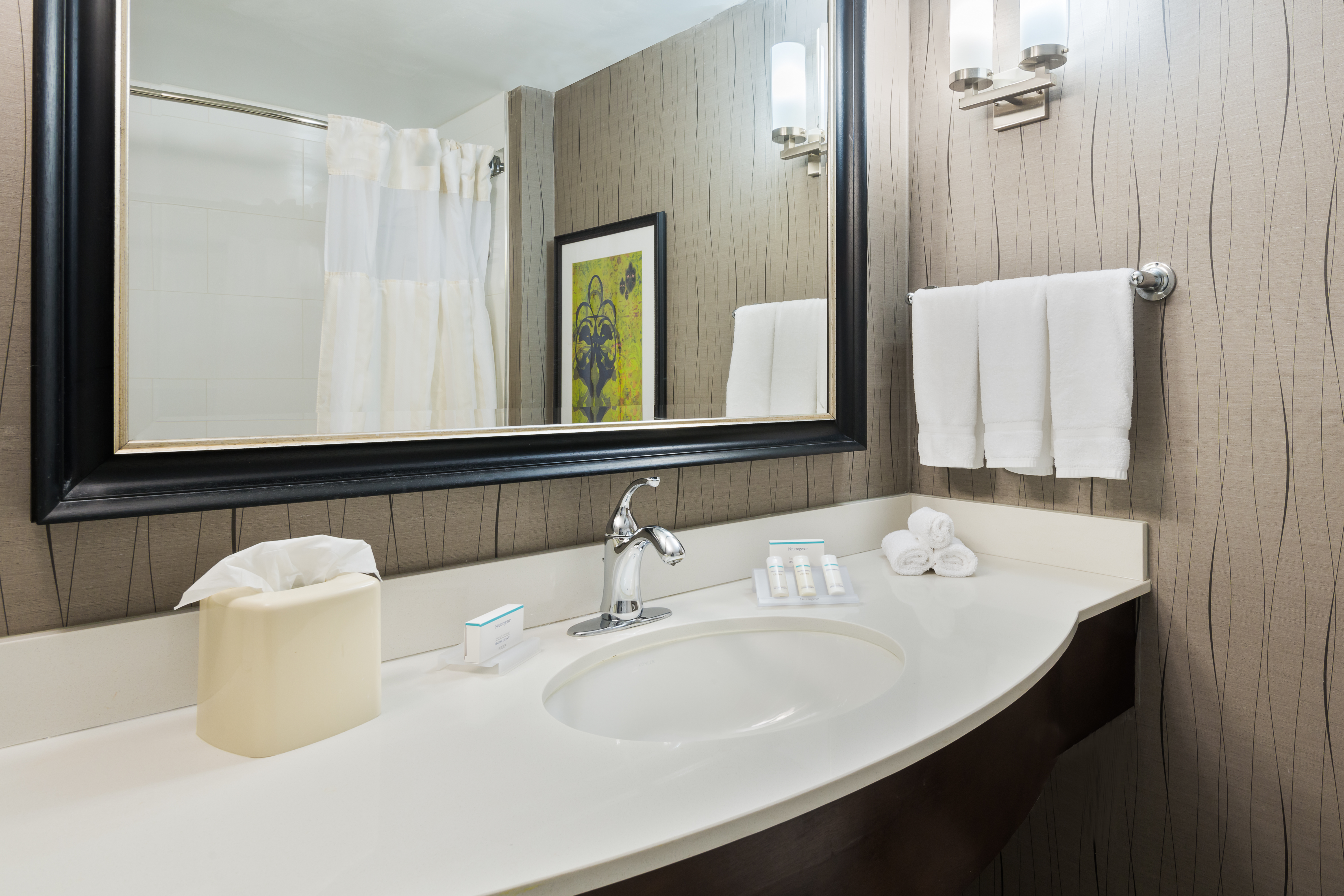 Shower Curtain and Wall Art Reflected in Large Vanity Mirror, Sink, Fresh Towels, and Toiletries in Standard Bathroom 