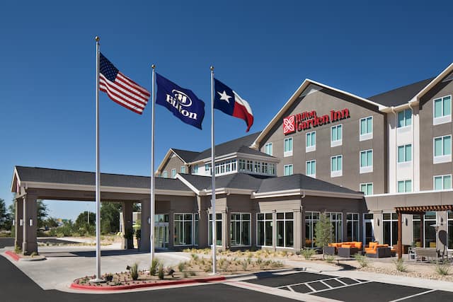 Side View of Hotel Exterior, Circle Driveway, Signage, Flagpoles, Landscaping, and Parking Lot on Sunny Day