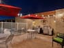 Two Tables With Red Umbrellas and Chairs, Soft Seating, and Two Barbecue Grills on Illuminated Outdoor Patio at Night