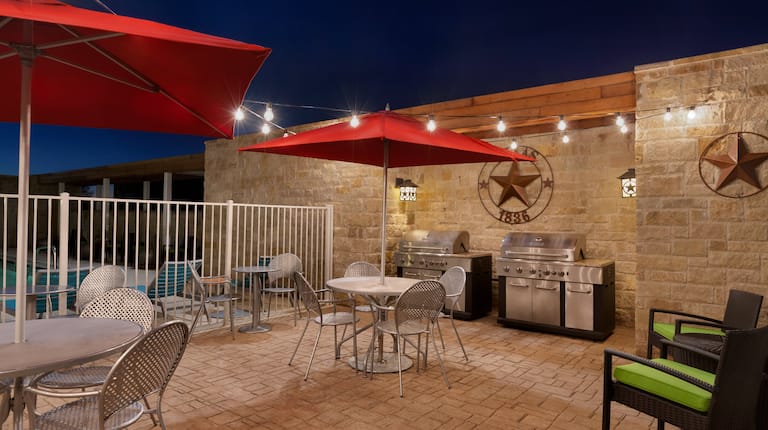 Two Tables With Red Umbrellas and Chairs, Soft Seating, and Two Barbecue Grills on Illuminated Outdoor Patio at Night