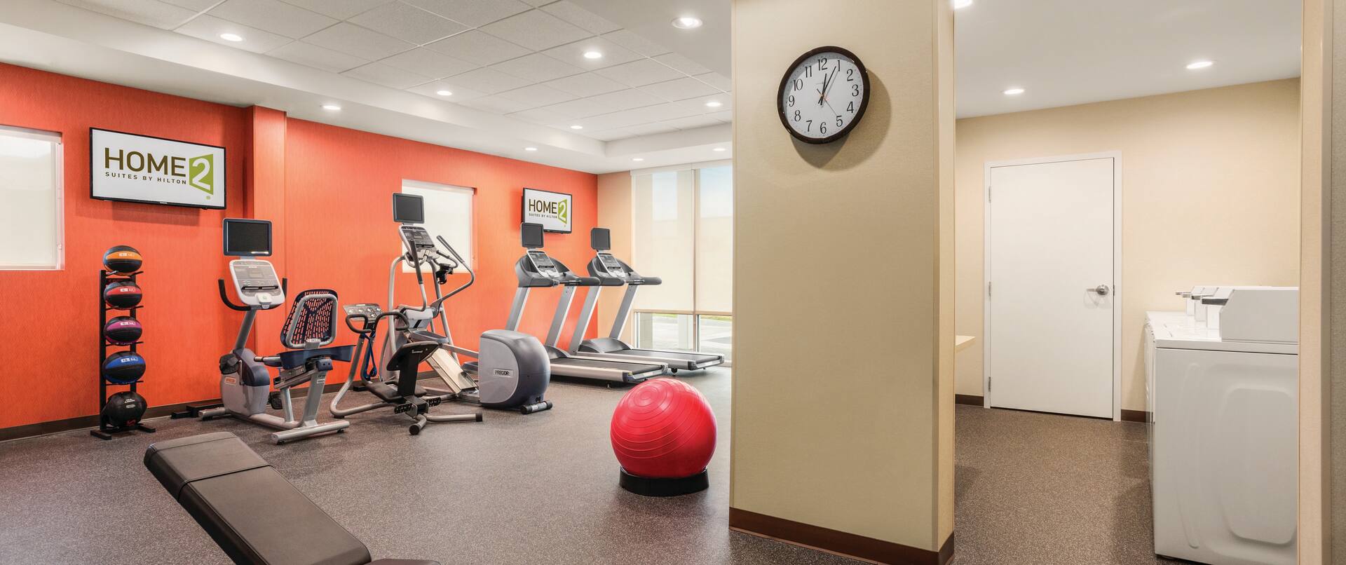 Wall Clock by Door Opening to Laundry Area, Weight Bench, Weight Balls, Cardio Equipment Facing TVs, and Red Exercise Ball in Fitness Center of Spin2 Cycle