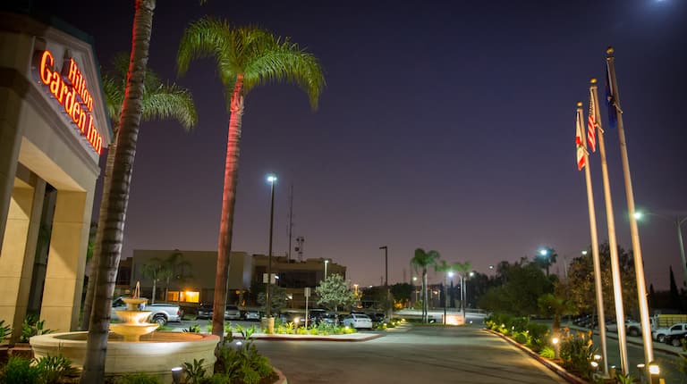 Partial View of Hotel Exterior, Signage, Fountain, Landscaping, Circle Driveway, Guest Cars on Parking Lot, and Flag Poles Illuminated at Night