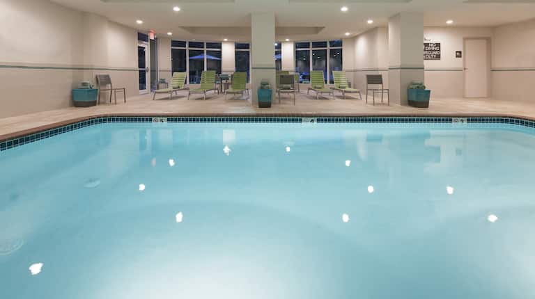 Chairs, Loungers, and Towel Stations by Window of Indoor Pool Illuminated at NIght
