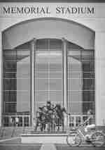 Seorang wanita bersepeda melewati stadion peringatan dengan patung pemain sepak bola di depannya 