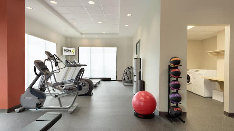  Spin2Cycle Fitness Area With Weight Bench, Cardio Equipment Facing Windows, TV in Corner, Free Weights, Large Mirror, Water Cooler, Red Exercise Ball, Weight Balls, and Open Doorway to Laundry Room