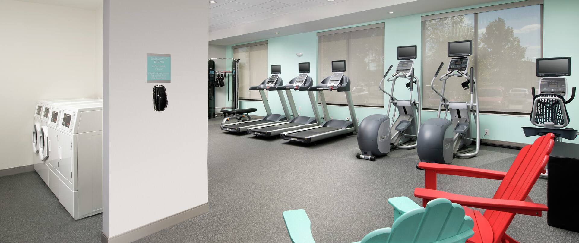 Spin2Cycle Fitness Area With Cardio Equipment, Lounge Seating, and Open Doorway to Laundry Room With Coin Operated Washing and Drying Machines