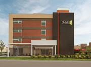 Modern Home2 Suites hotel exterior featuring pristine landscaping, patio with umbrellas and furniture, and bright blue sky.