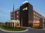 Modern Home2 Suites hotel exterior featuring porte cochere, American flag, and glowing patio.