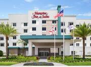 Welcoming Hampton Inn hotel exterior featuring palm trees, American flag, and bright blue sky.