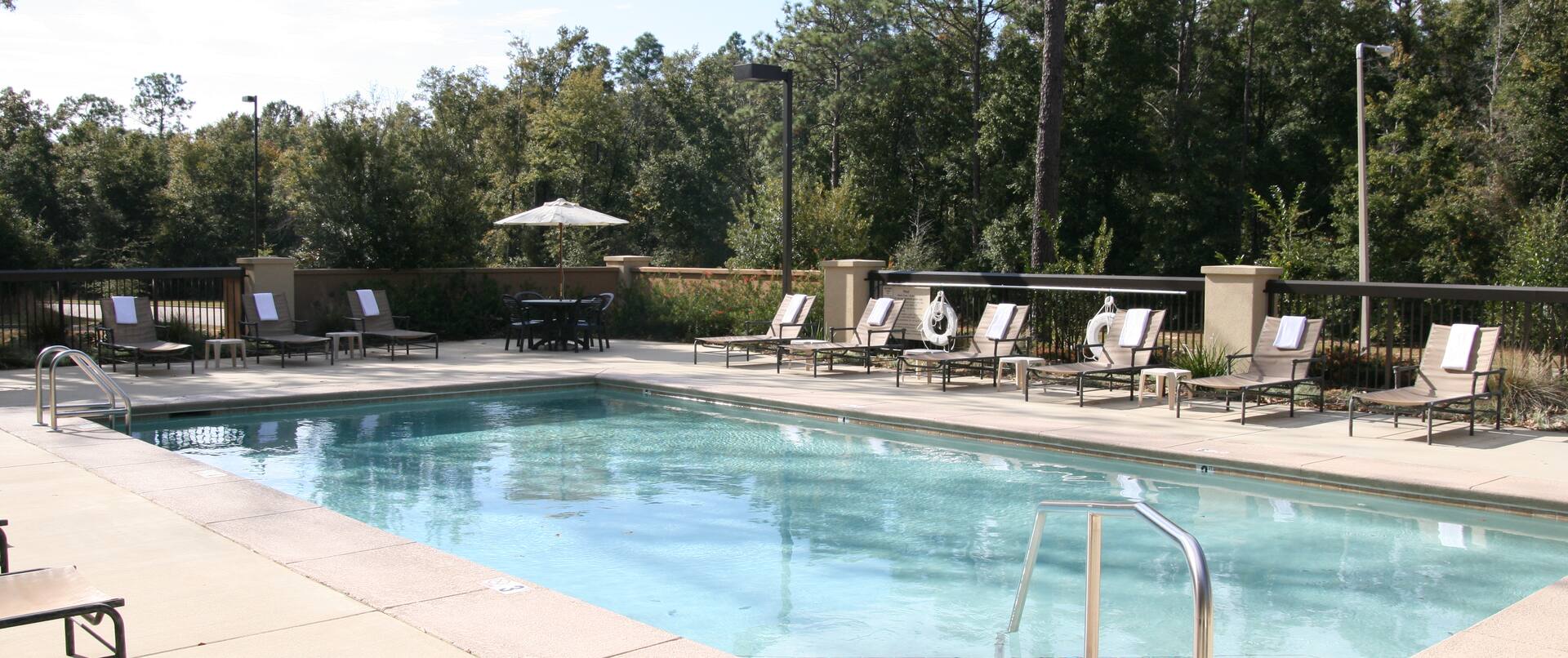 Daytime View of Outdoor Pool Surrounded by Trees, Table With Umbrella, Chairs, and Loungers on a Sunny Day