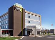 Modern Home2 Suites Hotel Exterior featuring porte cochere, beautiful landscaping, and bright blue sky.