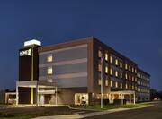 Modern Home2 Suites hotel exterior featuring glowing guestroom windows, outdoor patio, and dusk sky.