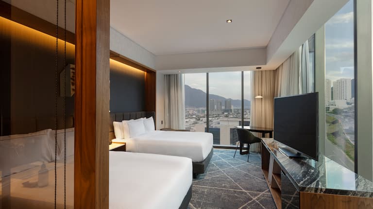 Guest room with two queen-sized beds, TV and floor-to-ceiling windows with views of mountains and city