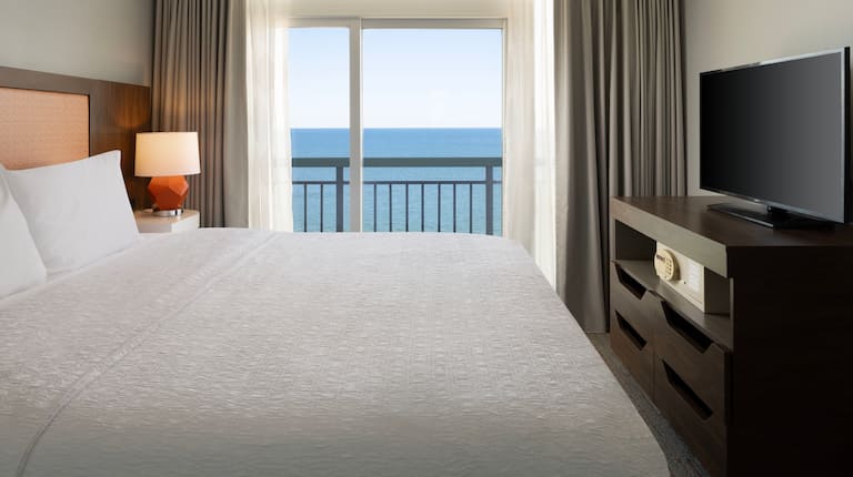 King bed in the suite bedroom with a wall of glass including a sliding glass door to balcony with views of the ocean.