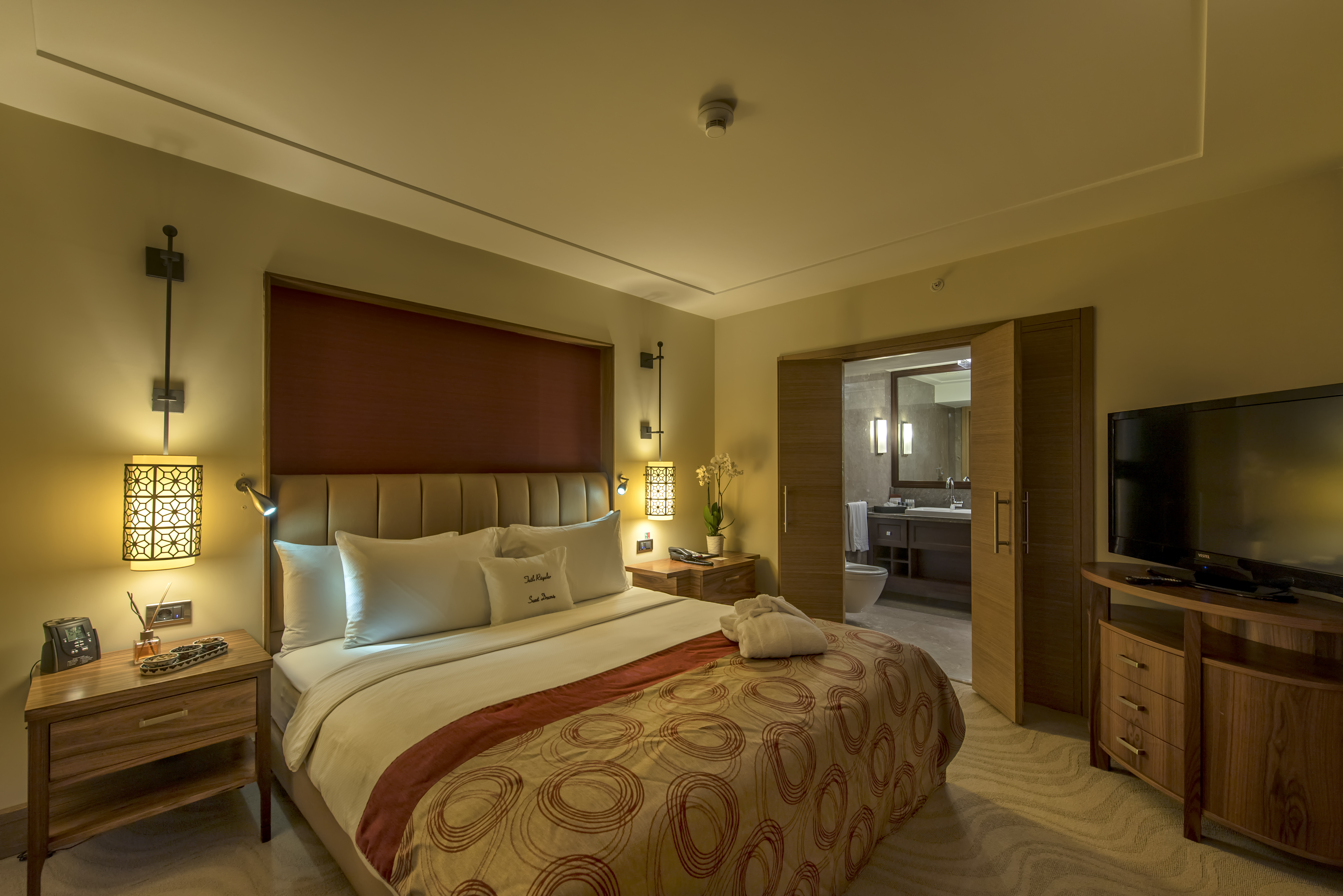 Bed, Illuminated Lamps Above Bedside Tables, Open Doorway to Bathroom, Dresser and TV in Suite 