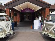 Two Gold and White Volkswagen Vans Decorated With White Ribbons and Flowers Parked by Wedding Entrance