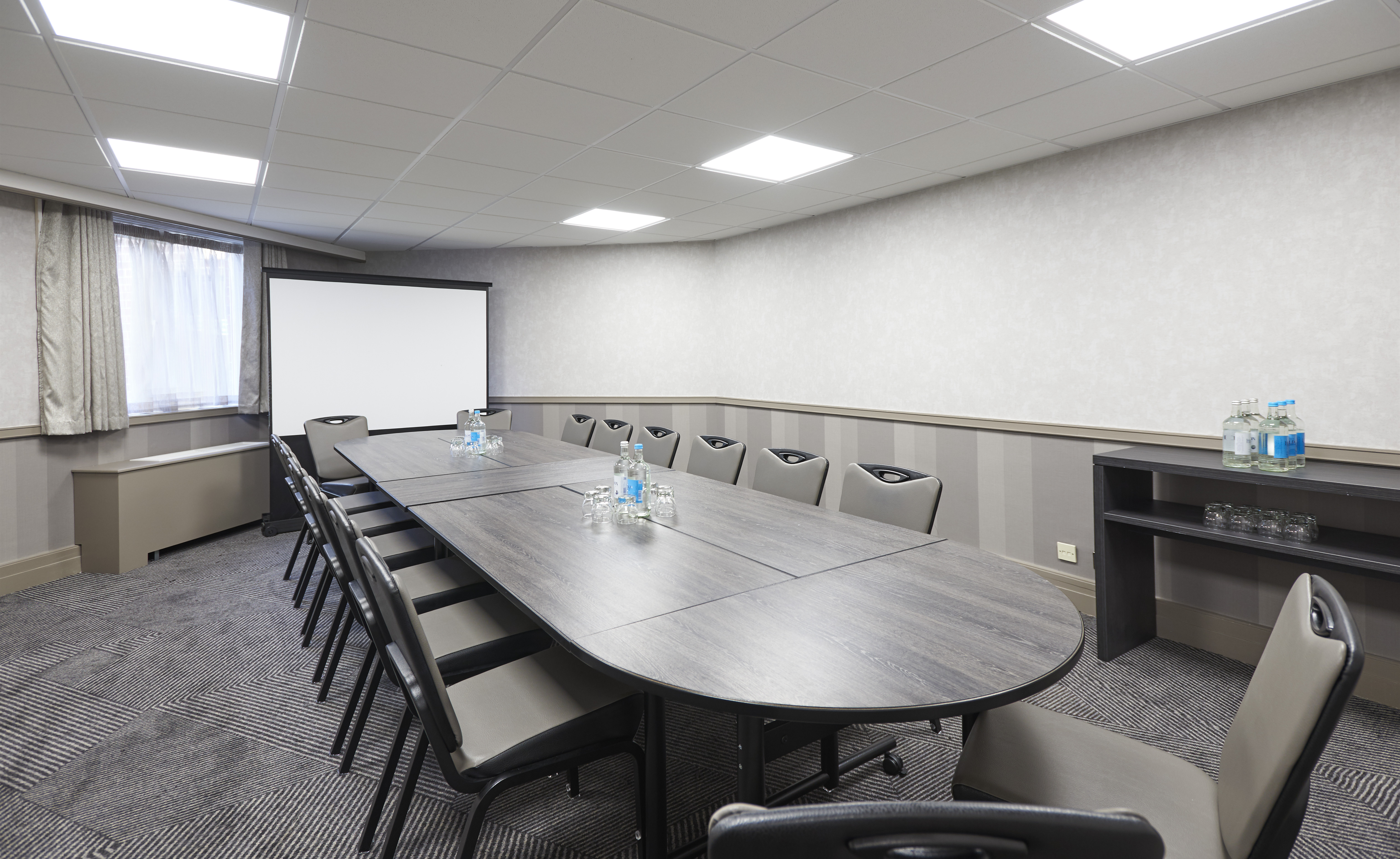 Angled View of Window With Sheer Drapes, Presentation Screen, Beverage Station, and Seating For 16 At Boardroom Table With Water Bottles and Drinking Glasses in Hermitage Suite