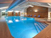 Illuminated Indoor Pool and Whirlpool With Lounge Chairs, Glass Door, and Large Windows With View into Fitness Center