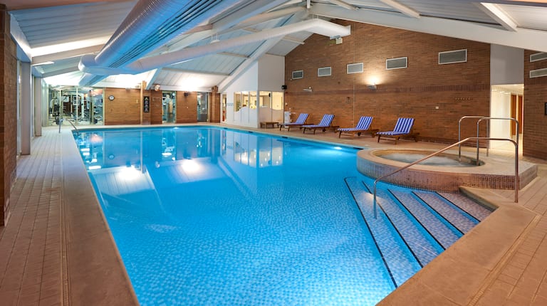 Illuminated Indoor Pool and Whirlpool With Lounge Chairs, Glass Door, and Large Windows With View into Fitness Center