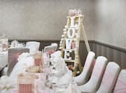 Detailed View of White Chairs With Pink Bows, Place Settings, Wine Glasses on Head Table With White Linens, and Small Ladder in Corner With White Flowers and Illuminated "Love" Light Set Up For Wedding Reception