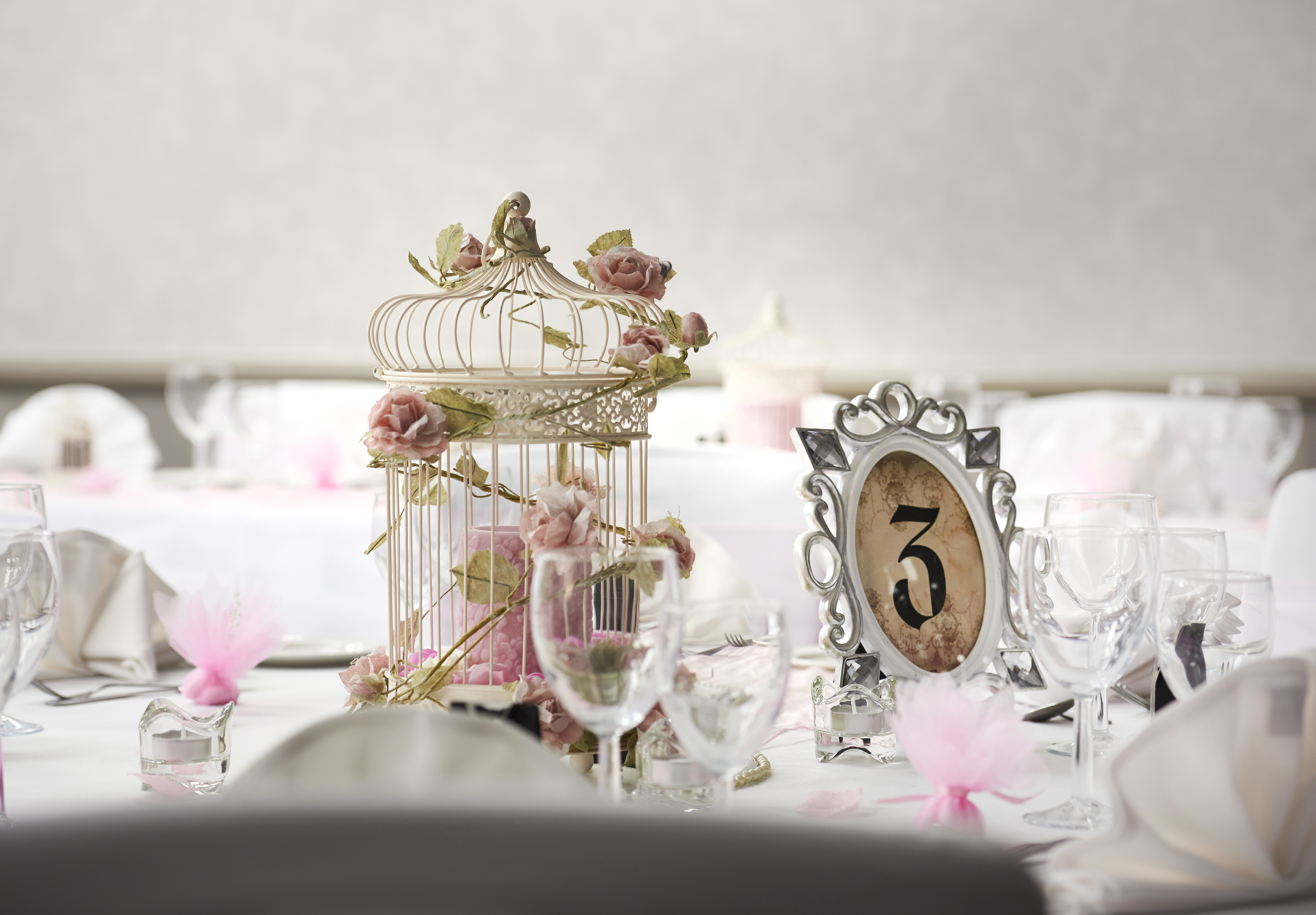 Place Settings, Wine Glasses, Framed Table Number, Bird Cage, and Wedding Favors on Round Tables With White Linens Decorated for Wedding Reception