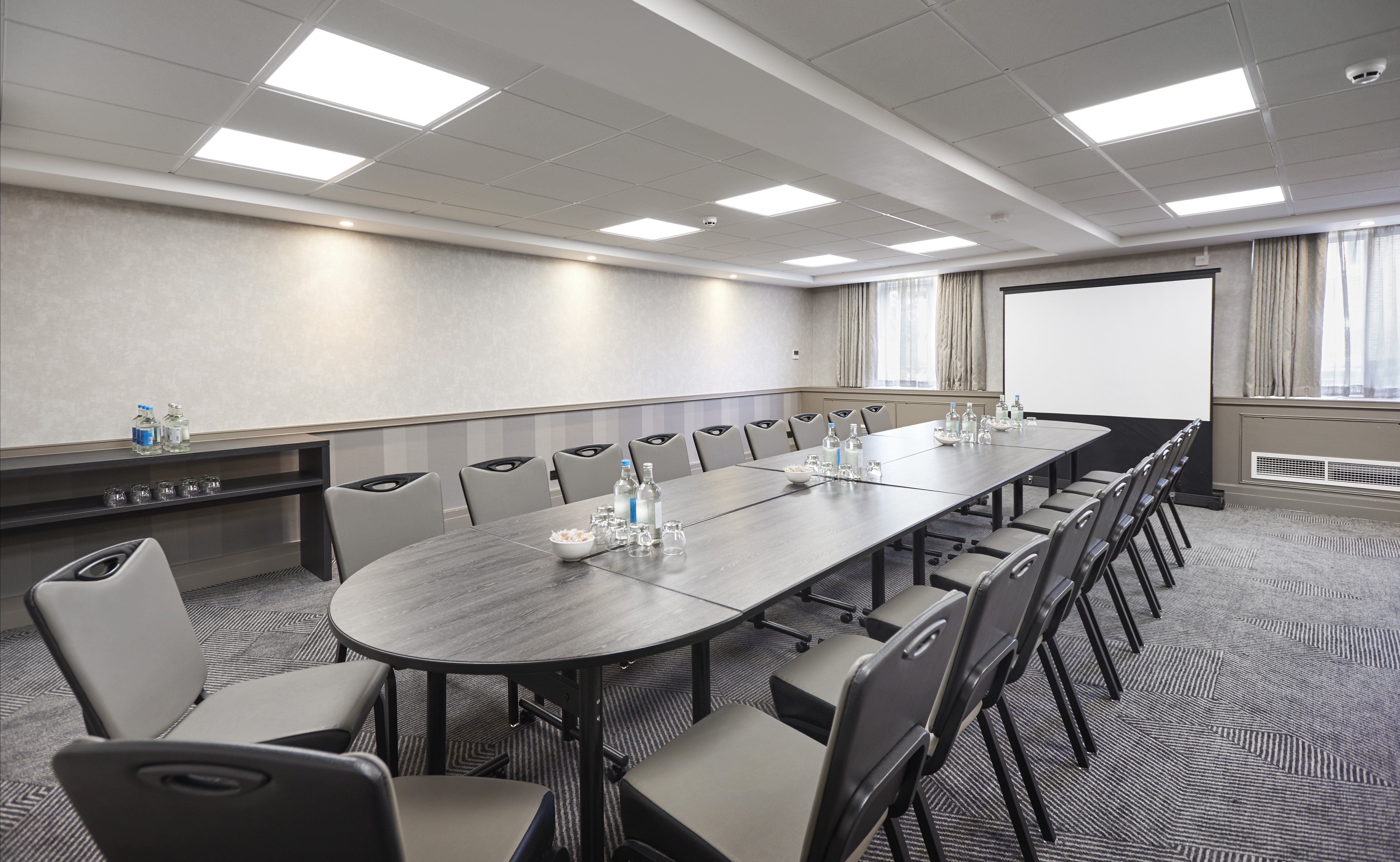Angled View of Beverage Station, Windows With Sheer Drapes, Presentation Screen, and Seating For 20 At Boardroom Table With Water Bottles and Drinking Glasses in Enbourne Meeting Room
