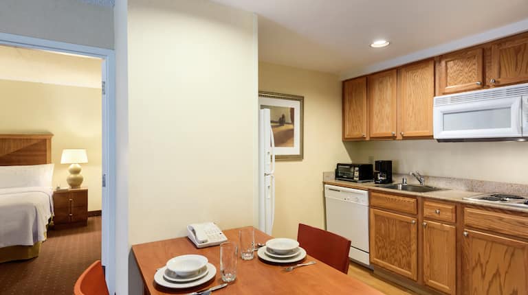 Kitchen With Fridge, Dishwasher, Toaster, Coffee Maker, Sink, and Microwave Over Stovetop,  Seating for Two at Dining Table, and Open Doorway to Bedroom 