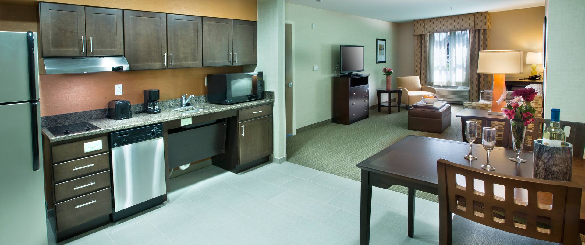 Suite Kitchen With Wall Art, Fridge, Wood Cabinets, Microwave Over Stovetop, Sink, Dishwasher, Coffee Maker, and Dining Table With Seating for Two