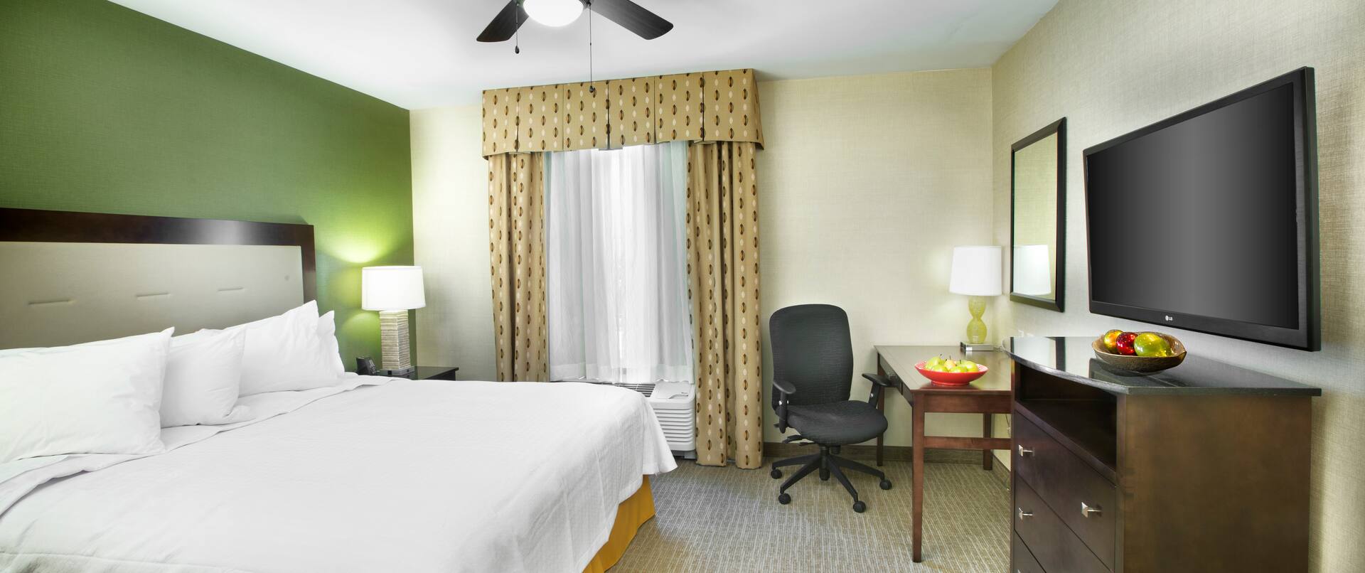 King Bed and Bedside Table With Lamp, Ceiling Fan, Work Desk by Window, and TV in Suite Guest Room