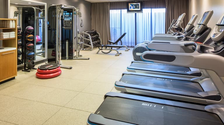Fitness Center With Cardio Equipment, Towel Station, Aerobic Stepper, Weight Balls, Wall Mirror, Weight Machine, Free Weights, Weight Bench, TV, and Window With Sheer Drapes