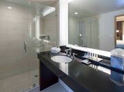 Shower With Glass Doors, View of Bedroom Reflected in Large Vanity Mirror, Sink, Toiletries, and Fresh Towels in Suite Bathroom
