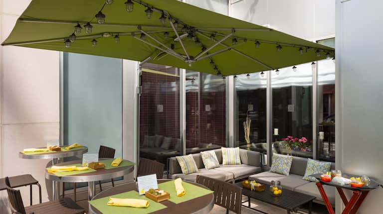 Daytime View of Dining Tables Under Green Umbrella and Lounge Seating By Hotel Exterior